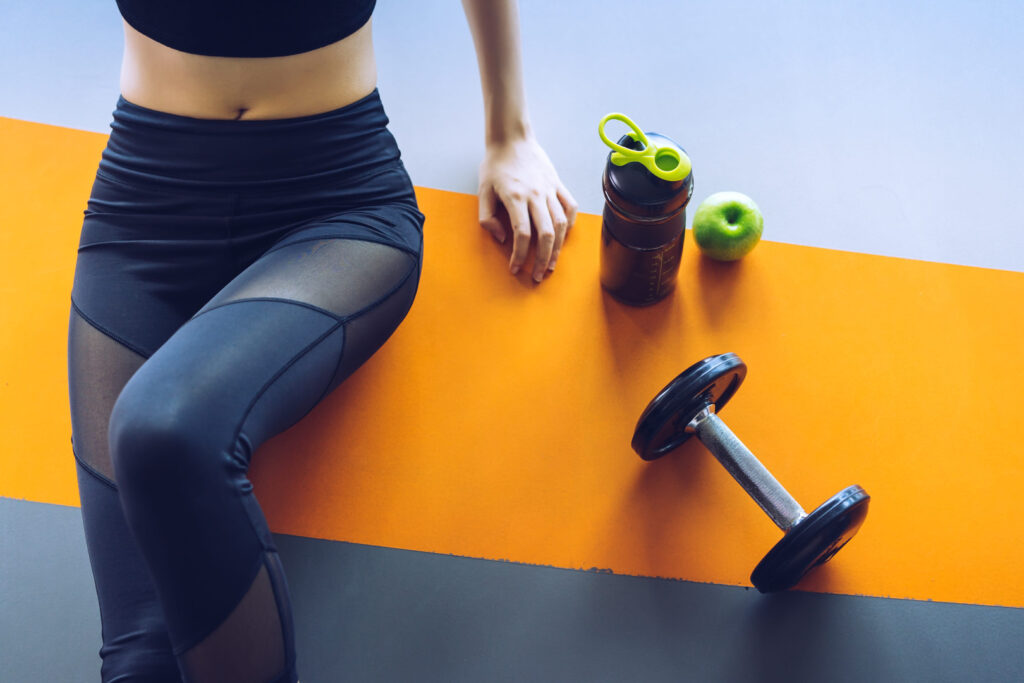 Edzés után pihenő nő az edzőteremben almával, súlyzóval és fehérjeturmixszal felülnézetből, a hirtelen fogyás helyett egészséges életmódra fókuszálva