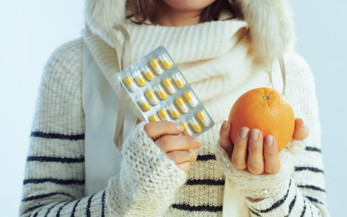 Immunrendszer-erősítés télen: meleg pulóvert, sálat és fülvédős sapkát viselő nő narancsot és vitaminkapszulákat tart világoskék háttér előtt.