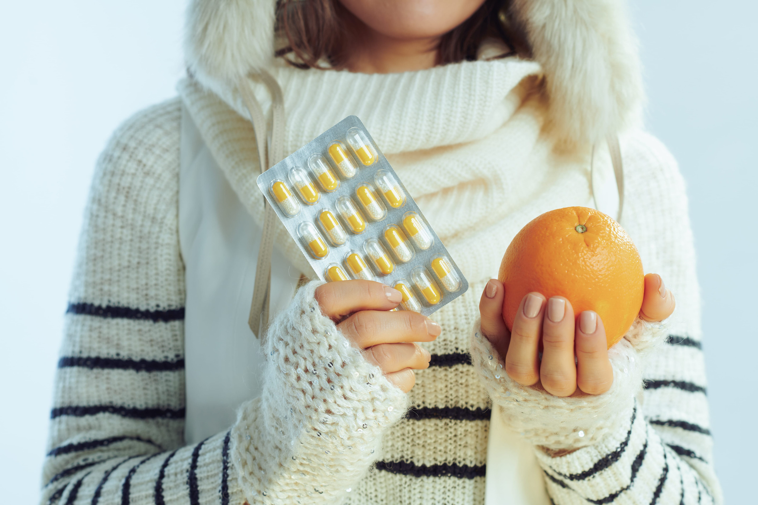 Immunrendszer-erősítés télen: meleg pulóvert, sálat és fülvédős sapkát viselő nő narancsot és vitaminkapszulákat tart világoskék háttér előtt.